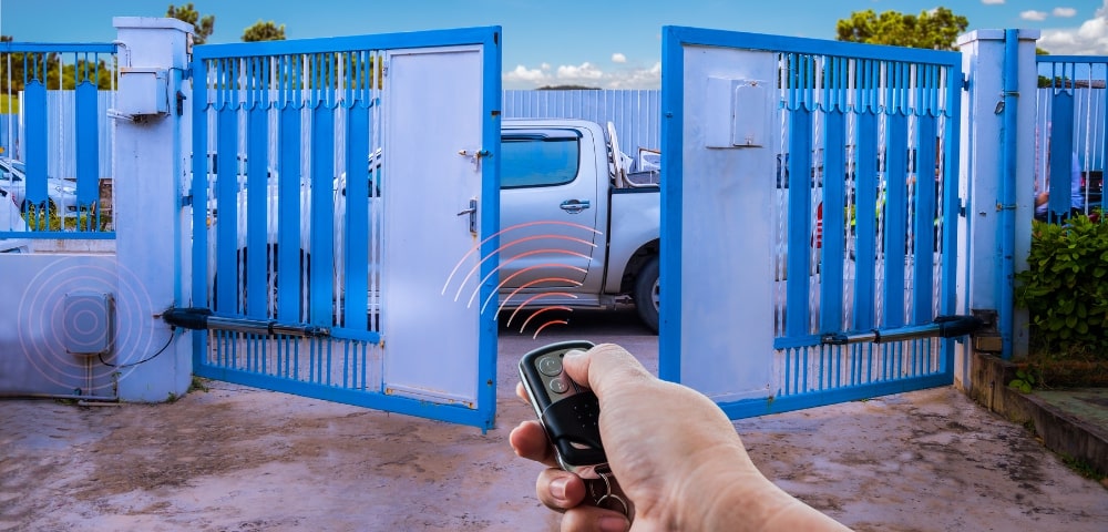 Mână cu telecomandă deschide o barieră automată la intrarea unei parcări rezidențiale, braț metalic și motor acționare vizibile