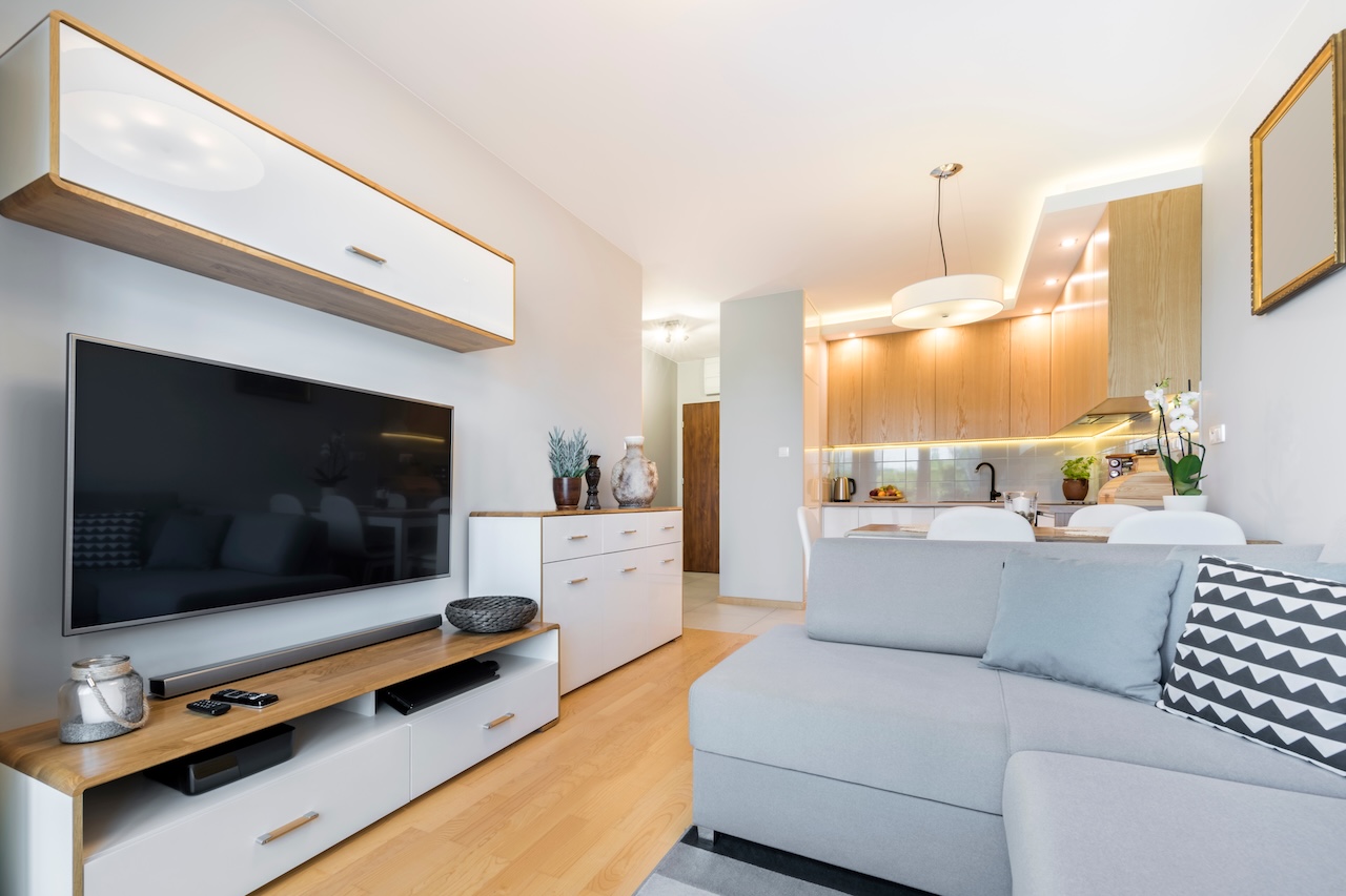 Living open-space amenajat cu o canapea de colț gri deschis, comodă TV albă, mobilier din lemn și bucătărie deschisă.