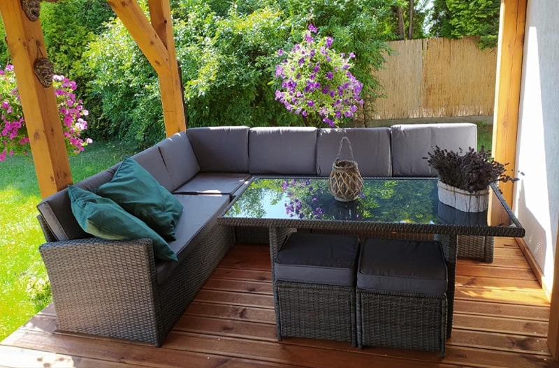 Mobilier de grădină din ratan sintetic, cu masă din sticlă și colțar cu perne gri, amenajate pe o terasă din lemn într-un spațiu verde.