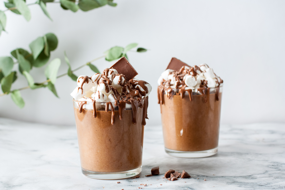 Ciocolată caldă de casă cu frișcă și topping de ciocolată, servită în pahare de sticlă pe masă din marmură.