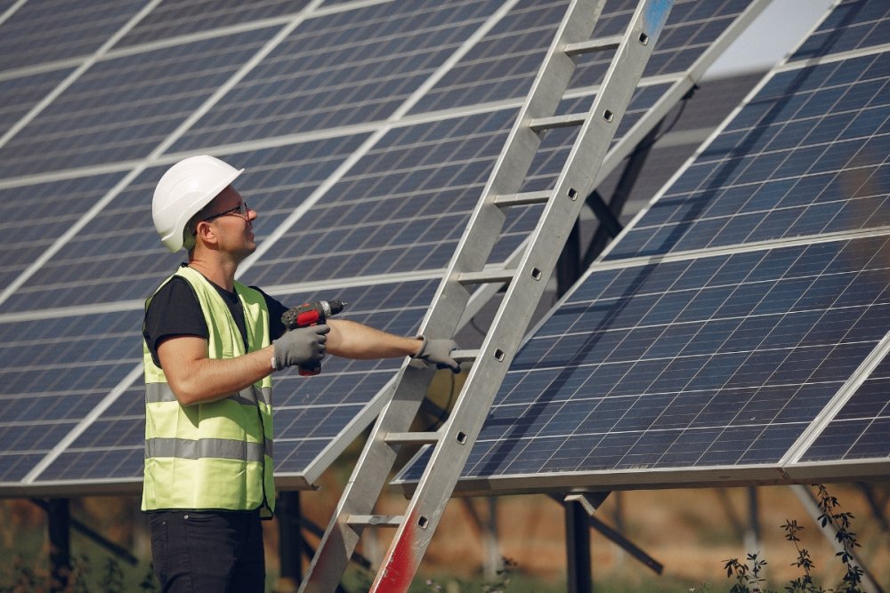 Tehnician cu echipament de protecție, lângă o scară și un sistem de panouri fotovoltaice montate pe structură metalică.