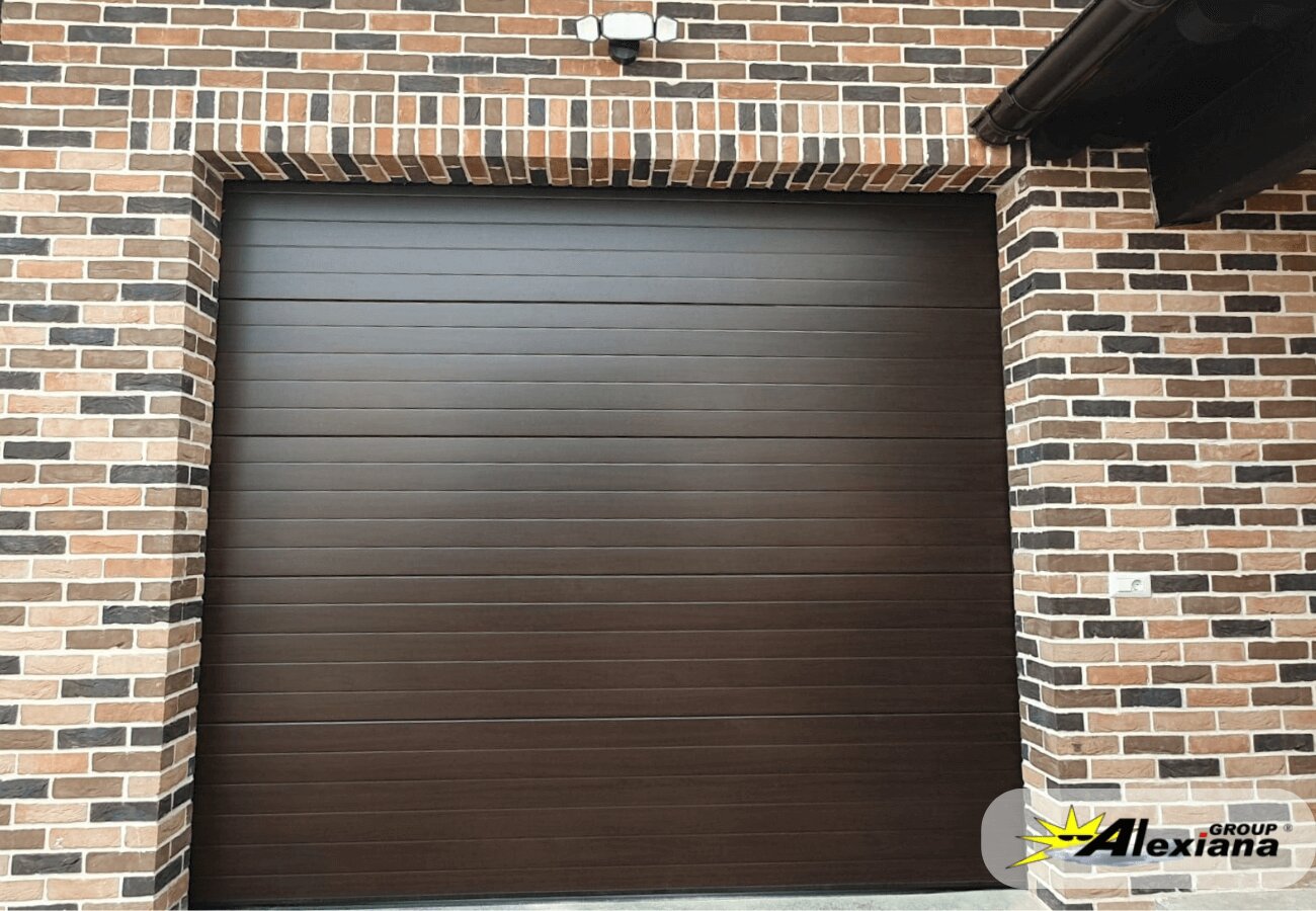 Ușă de garaj secțională modernă, finisaj maro închis, montată pe fațadă din cărămidă aparentă, soluție termoizolată Alexiana Group.
